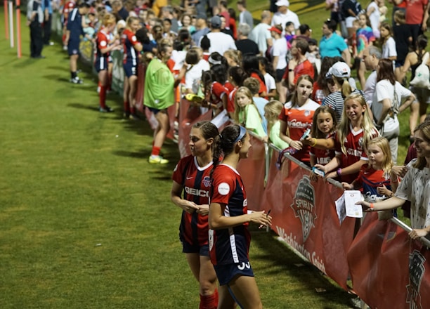 The image features a lively gathering of young soccer players and fans next to a field. Several female athletes in red and blue uniforms interact with an enthusiastic crowd standing behind a barrier. Some people in the crowd are holding objects, like pens and paper, possibly for autographs.