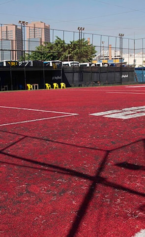 A sports court with a red playing surface and white lines, surrounded by a black mesh fence. In the background, tall buildings are visible, along with some trees and a few yellow chairs positioned near the fence.
